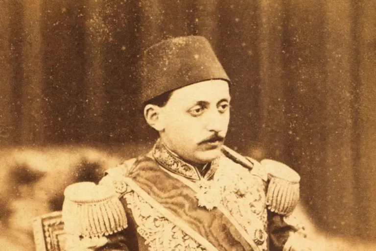 Sepia photograph of a young man with a moustache in a fez, seen from the chest up, in a richly embroidered uniform with a sash and fringed epaulettes; his face is turned to the left