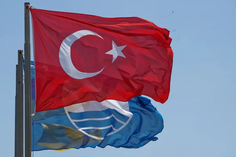 A red Turkish flag flying in front of a blue flag with the white wave sign, on tall poles against a clear sky