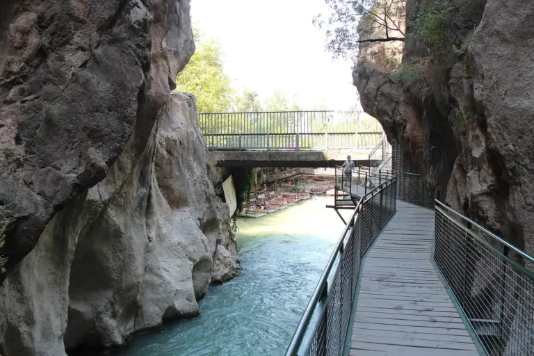 A wooden walkway with mesh railings runs along a rock wall above a turquoise river in a narrow gorge, with a road bridge across the river ahead and one person standing on the walkway in the distance