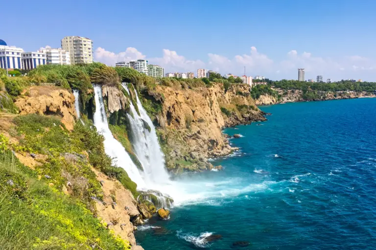 Water dropping over a golden cliff straight into the blue sea, with a line of white apartment blocks along the cliff top and a green slope in the foreground