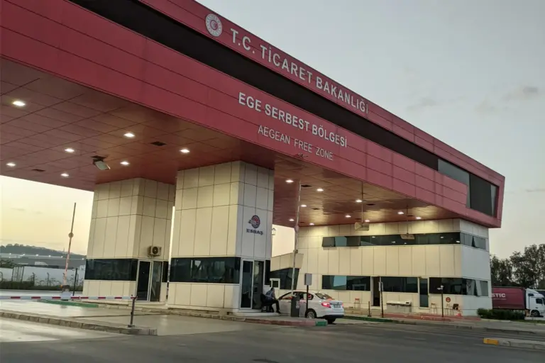 The red entrance building of the Aegean Free Zone in İzmir, with the Ministry of Trade's name and «Ege Serbest Bölgesi, Aegean Free Zone» on its front, a barrier, a car at the booth and a lorry beyond