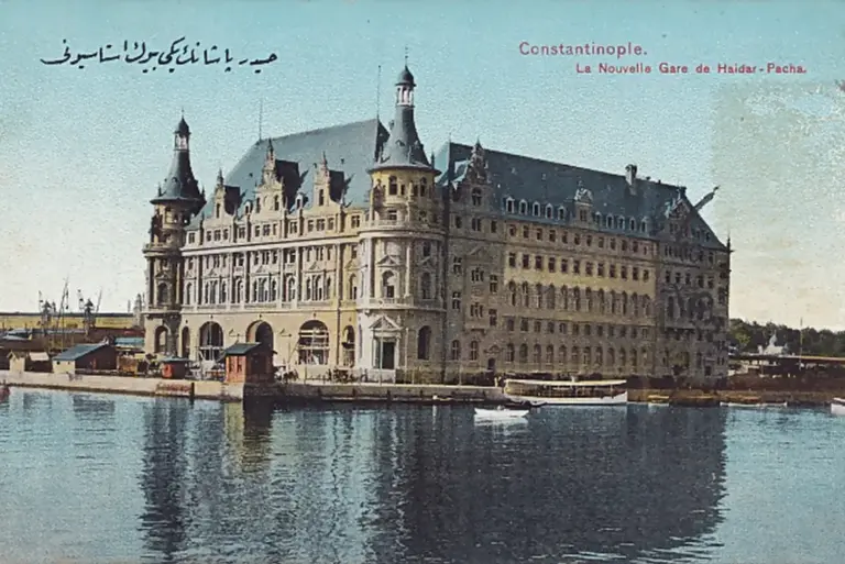 Colour postcard of Haydarpaşa station seen across the water: a long stone building with a steep slate roof, round corner towers with pointed caps and rows of arched windows, a small passenger boat at the quay, and the printed words «Constantinople. La Nouvelle Gare de Haidar-Pacha»