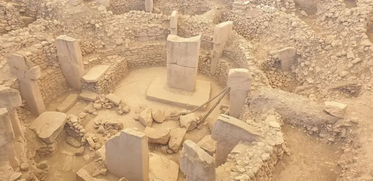 An aerial view of an oval stone enclosure in a dug pit: pillars stand in its wall and two taller pillars stand in the middle, with steel supports beside them