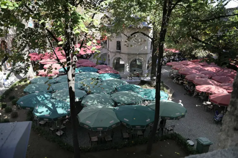 The courtyard of Koza Han in Bursa seen from an upper arcade: café tables under green and red umbrellas round the small domed prayer room, with plane trees overhead