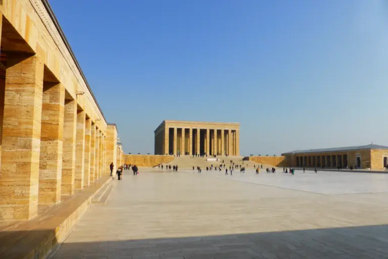 A wide stone plaza under a clear blue sky, with a long yellow-stone colonnade on the left and the columned Hall of Honour far across the plaza