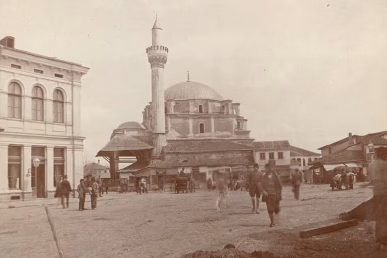 Photograph in sepia: the Banya Bashi mosque in Sofia with its domed prayer hall and a tall minaret, people and carts on the square in front and a building with arched windows on the left