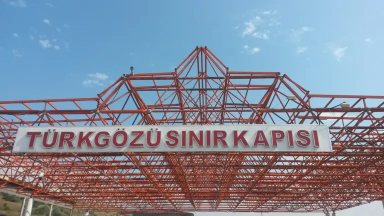 A white sign with red capital letters reading TÜRKGÖZÜ SINIR KAPISI on a red steel lattice roof under a blue sky