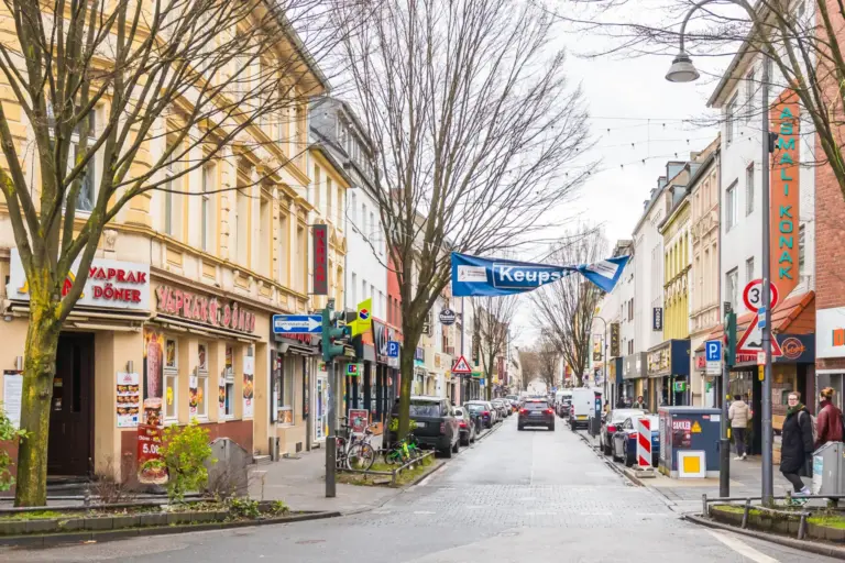A city street seen from its middle, with shops under bare trees, a banner across the road reading Keupstraße and tall shop signs in Turkish on both sides