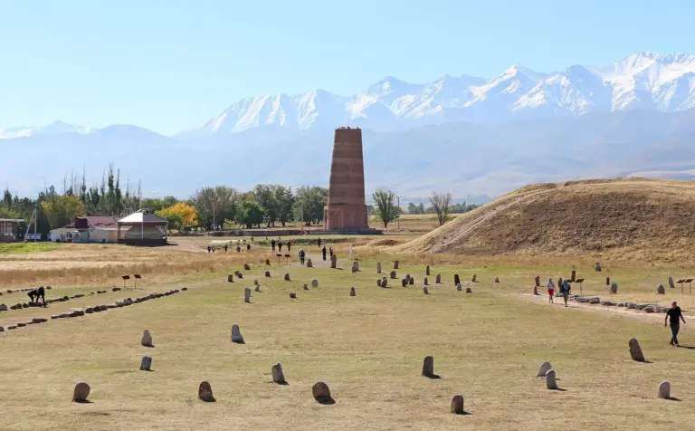 A tall round brick tower stands on a flat, dry grass plain with rows of small rounded standing stones in front of it and a long snow-covered mountain range behind; visitors walk between the stones