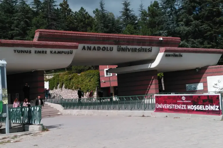The brick and white concrete gateway of the Yunus Emre campus under pine trees, with the signs ANADOLU ÜNİVERSİTESİ and YUNUS EMRE KAMPÜSÜ and a red welcome banner on the right