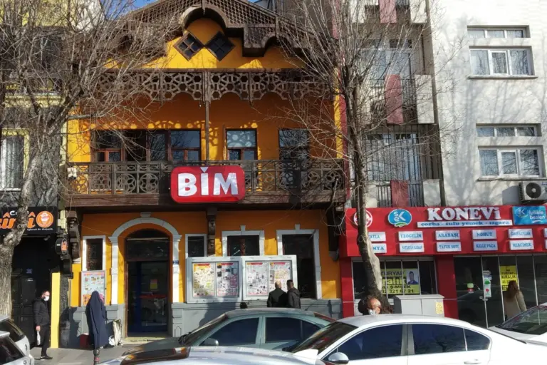 A yellow two-storey building with a carved wooden balcony on a street in Konya; a red BİM sign hangs on the balcony and brochure display cases stand beside the door
