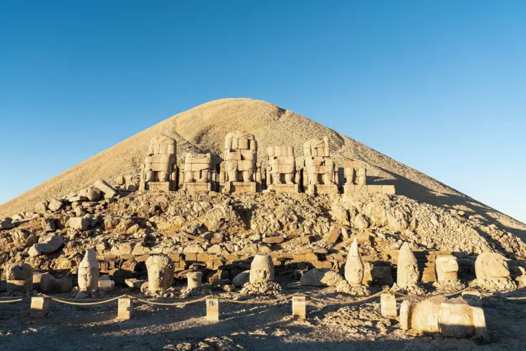 A row of seated stone thrones on a hilltop terrace in low light, with a cone of crushed stone rising behind them and stone heads on the ground in front