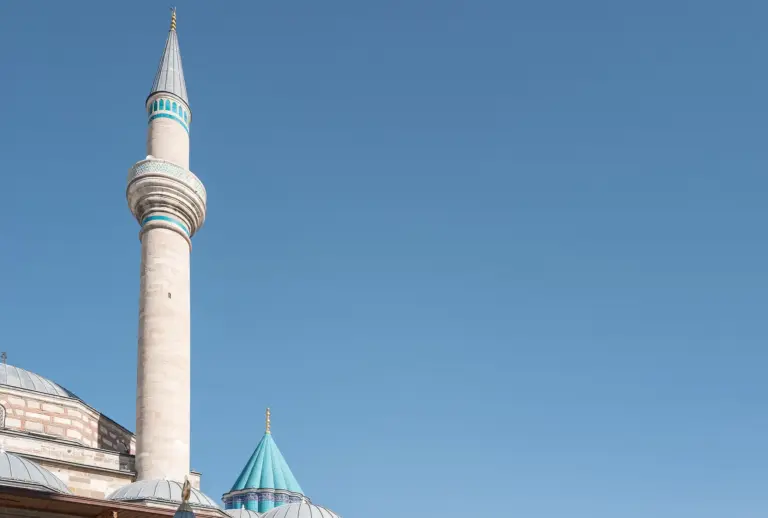 A tall stone minaret with turquoise bands, and beside it the pointed turquoise dome called the Green Dome, over grey lead domes under a clear blue sky