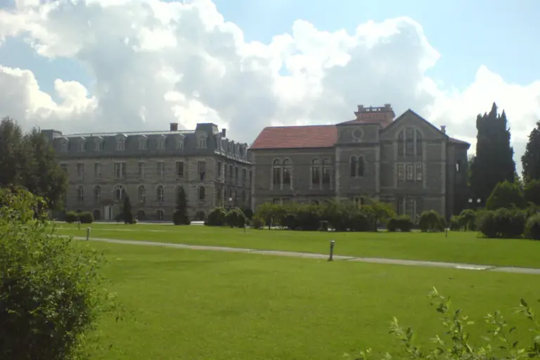 Two old stone buildings, one with a dark mansard roof and one with a red tiled roof, behind a wide lawn under tall white clouds: the South Campus of Boğaziçi University in Istanbul
