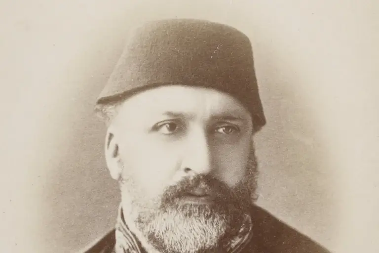 Sepia studio portrait of Sultan Abdülaziz in a fez: a heavy man with a full grey beard in a dark buttoned coat with a star-shaped decoration, looking slightly to the left