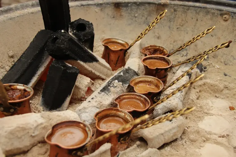 Small copper coffee pots with long twisted handles standing in pale ash beside black charcoal, each filled with frothy brown coffee