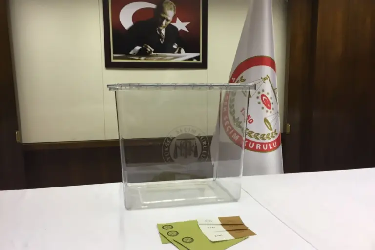 A transparent plastic ballot box on a white-covered table in front of a wooden wall with a framed portrait and a white flag with a round emblem, and a few ballot slips and olive-green envelopes on the table