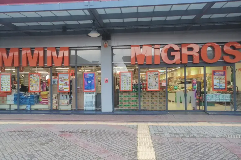 The glass front of a Migros store with large orange letters, MMM at the left and MiGROS at the right, and printed brochure sheets in the windows, seen from a paved square