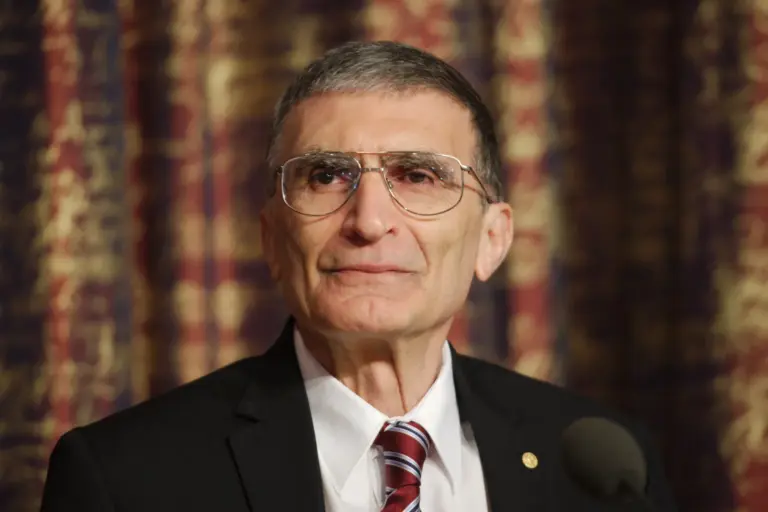 Portrait of Aziz Sancar: an older man with short grey hair and round glasses, in a dark jacket, white shirt and red-striped tie, looking at the camera with a slight smile in front of a dark red patterned curtain