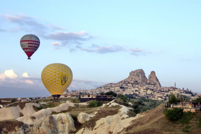 Two hot air balloons, one red and white and one yellow, in a pale morning sky beside a rock castle with houses on its slope