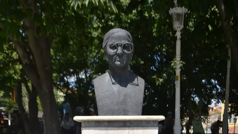 A dark grey bust of a person in round glasses with short hair, from the chest up, on a pale stone pedestal under green trees and beside a white lamp post