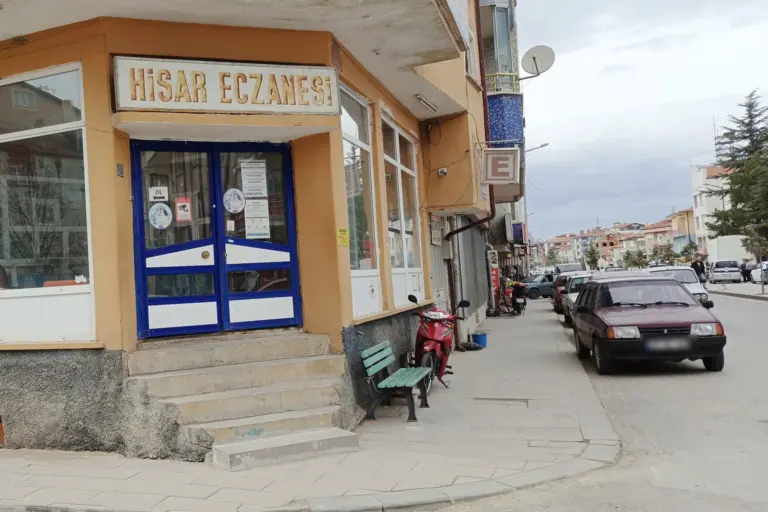 A corner pharmacy in Doğanhisar with a Hisar Eczanesi sign over a blue door, steps and a bench, and the red E pharmacy sign on the wall