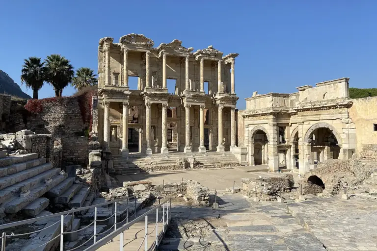 The pale stone front of the Library of Celsus at Ephesus: two storeys of columns, three doorways, three windows and a flight of steps, under a clear blue sky, with ruined walls on both sides