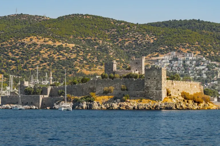 A castle of pale stone with towers and long walls on a rocky headland over deep blue sea, green hills and white houses behind it under a clear sky