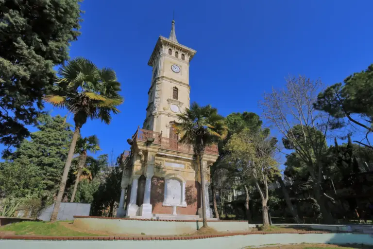 A tall clock tower of pale stone with a pointed roof, standing among palms and pines on a sunlit lawn under a deep blue sky