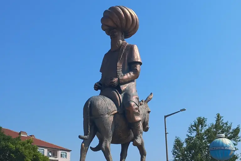 A statue of a bearded man in a very large turban sitting on a donkey against a clear blue sky, with a street lamp, a tree and a blue globe-shaped monument at the right edge