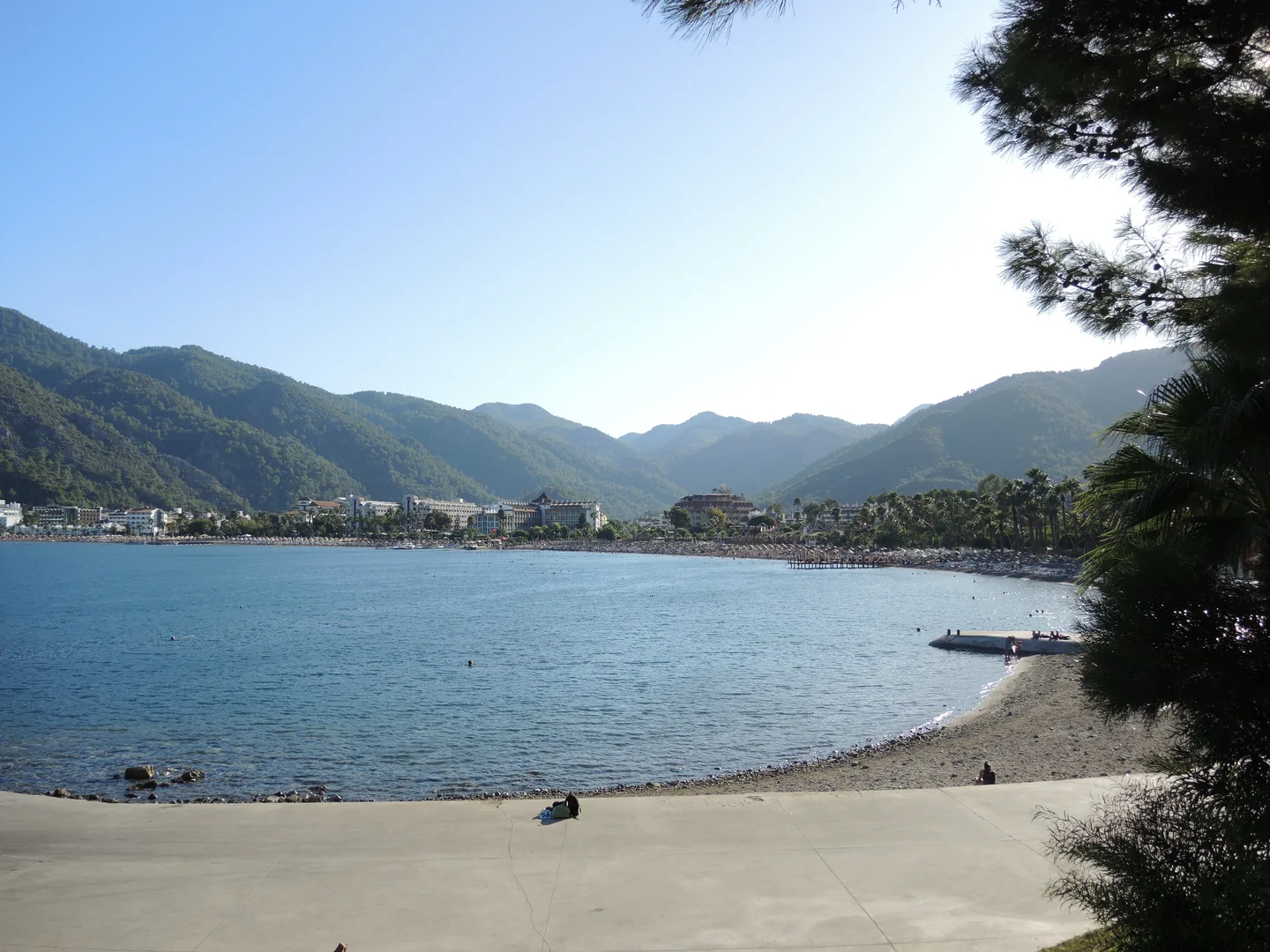 The curved beach and promenade at İçmeler, with resort buildings and wooded hills around the bay.