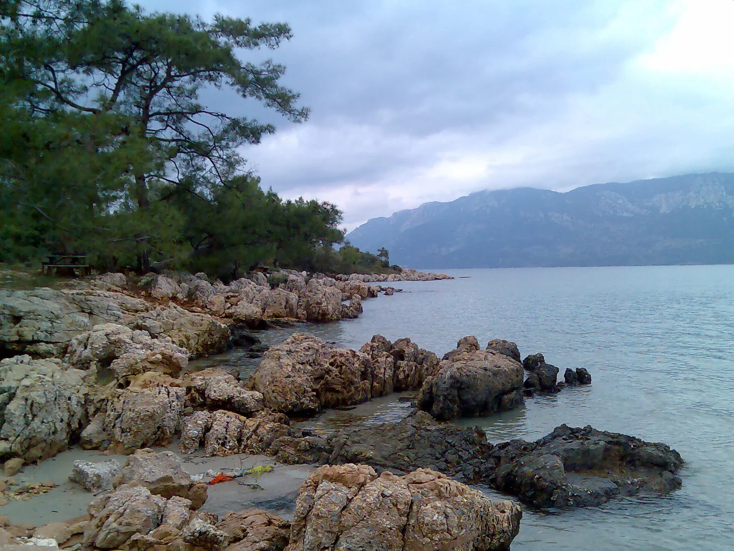 Pine trees and a rocky shoreline beside İncekum Beach near Çamlı, Marmaris, with clear water and mountains across the bay.
