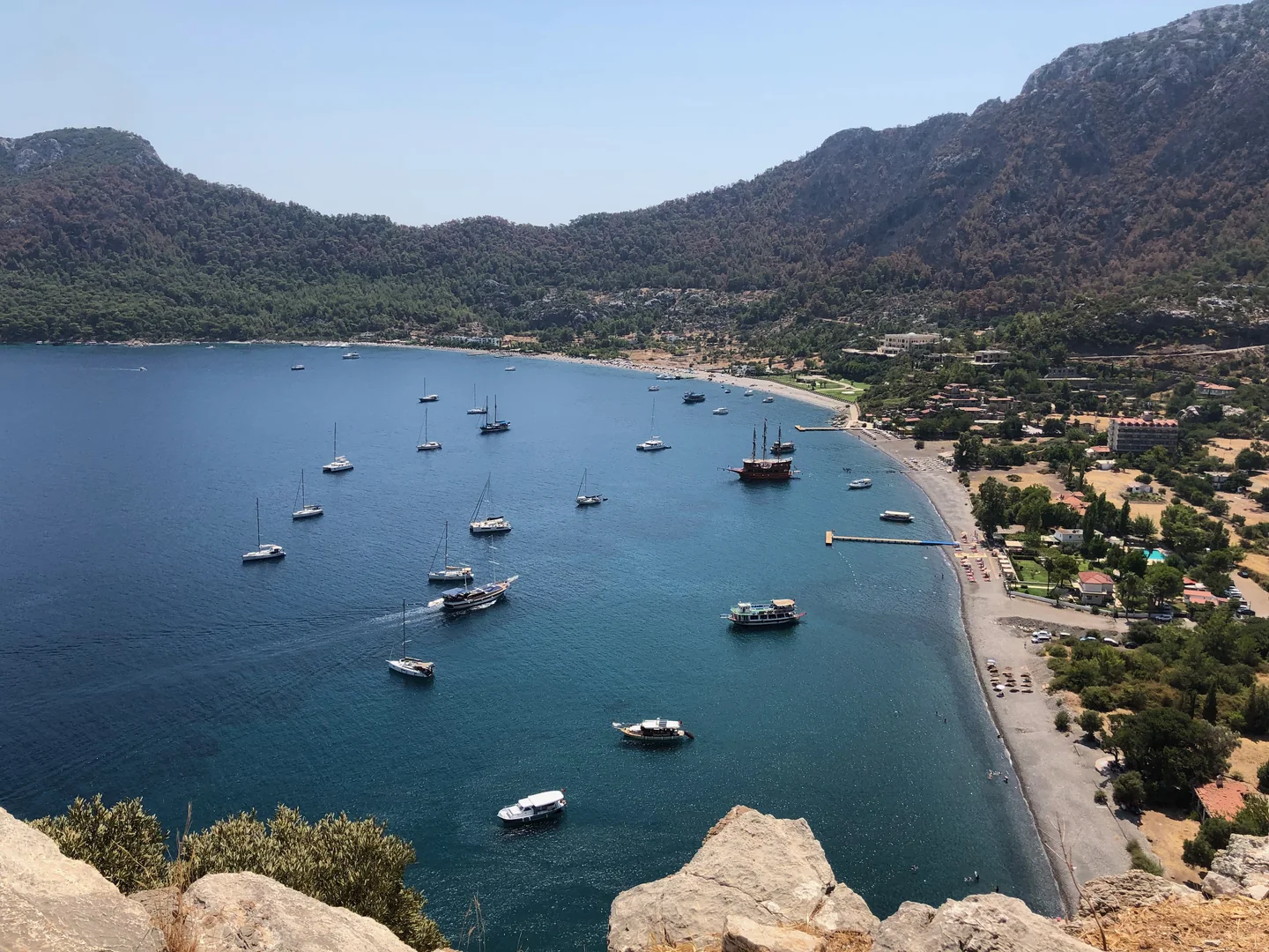 The long curved beach at Kumlubük, with boats in the bay and forested hills behind.
