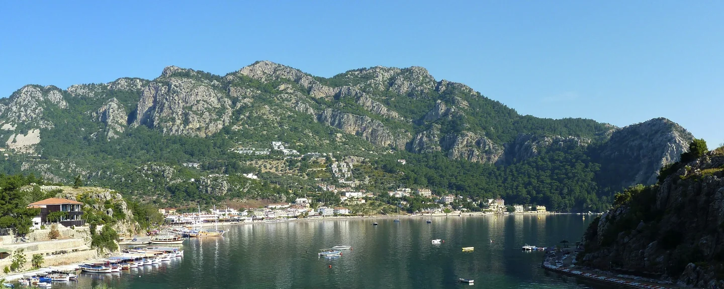 Panoramic view across Turunç bay toward its beach, waterfront village and steep hills.