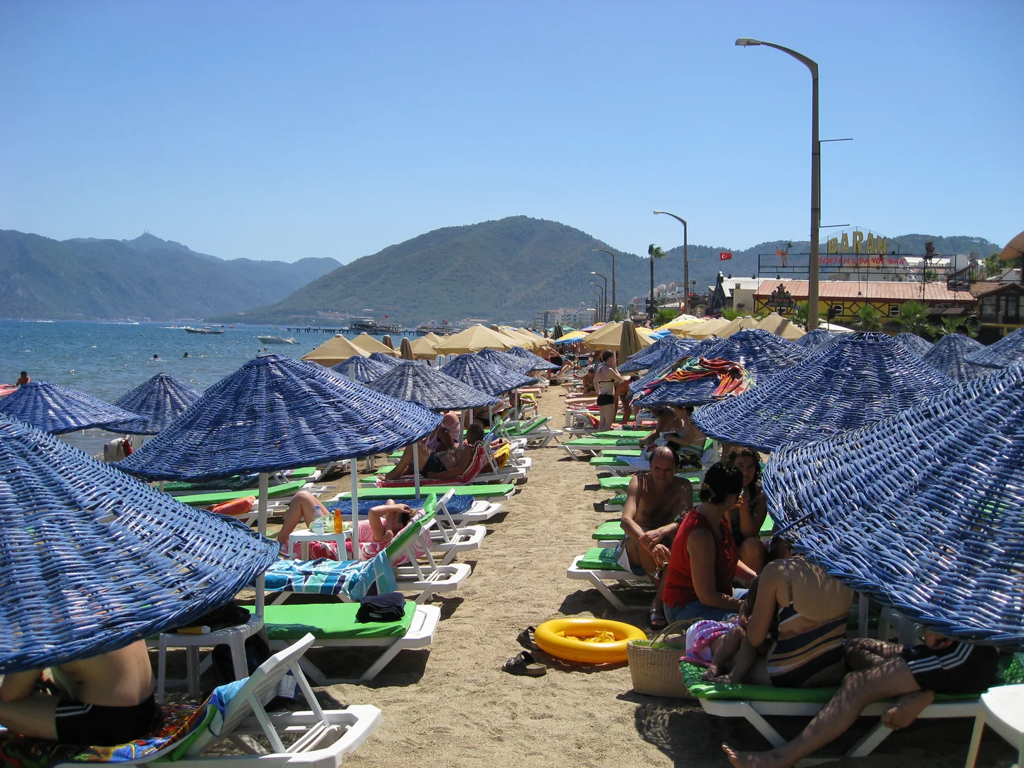 Rows of blue parasols and loungers on Uzunyalı beach near Yunus Hotel, with the Marmaris bay and hills beyond.