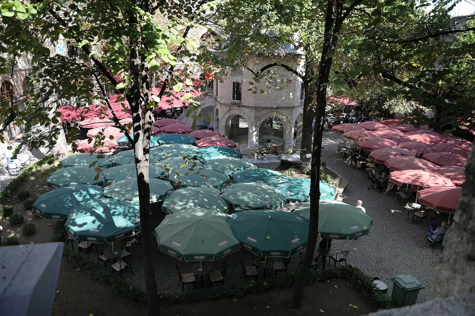 The courtyard of Koza Han in Bursa seen from an upper arcade: café tables under green and red umbrellas round the small domed prayer room, with plane trees overhead