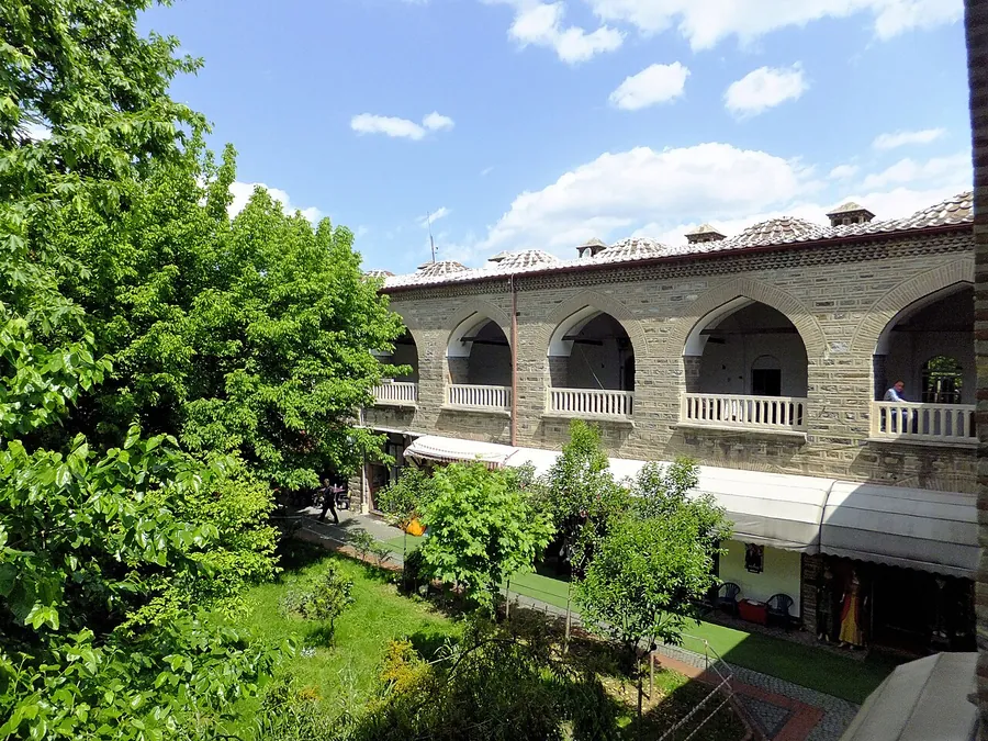 A courtyard garden in Fidan Han in Bursa, with trees in front of a two-floor stone arcade under a blue sky