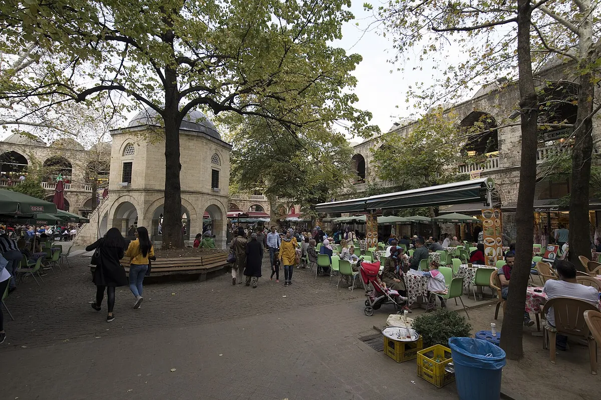 The courtyard of Koza Han in Bursa at street level: a small domed prayer room under plane trees, café tables and people, and the stone arcade behind