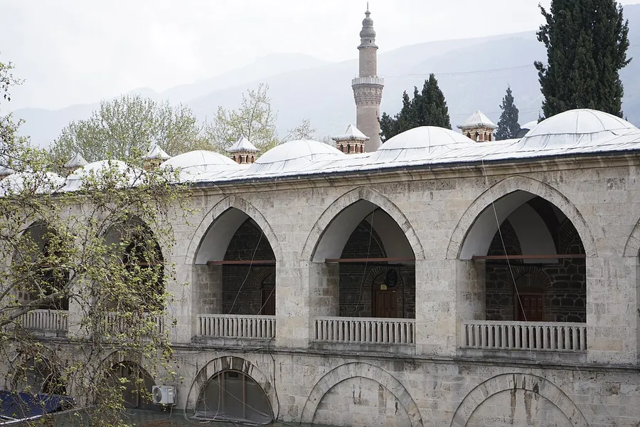 The two-floor stone arcade of Pirinç Han in Bursa, with pointed arches, balconies and domes behind it, and the Ulu Cami minaret above