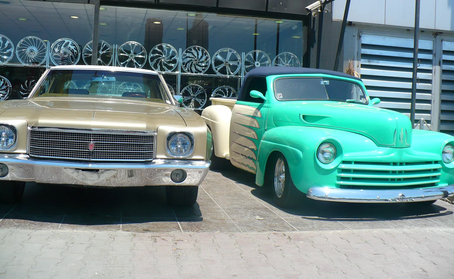 Two classic cars parked in front of a glass-fronted showroom with wheels on display: a gold coupé on the left and a green convertible pickup on the right