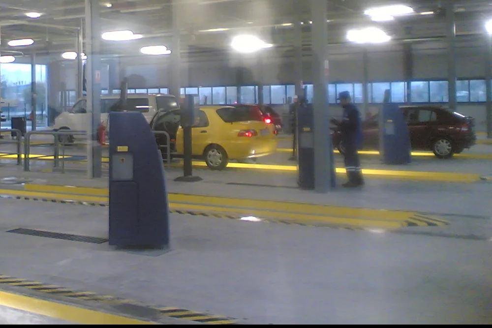 Cars on the lanes of a vehicle inspection station: a yellow hatchback and a dark saloon wait beside blue test posts while an inspector in overalls walks between them