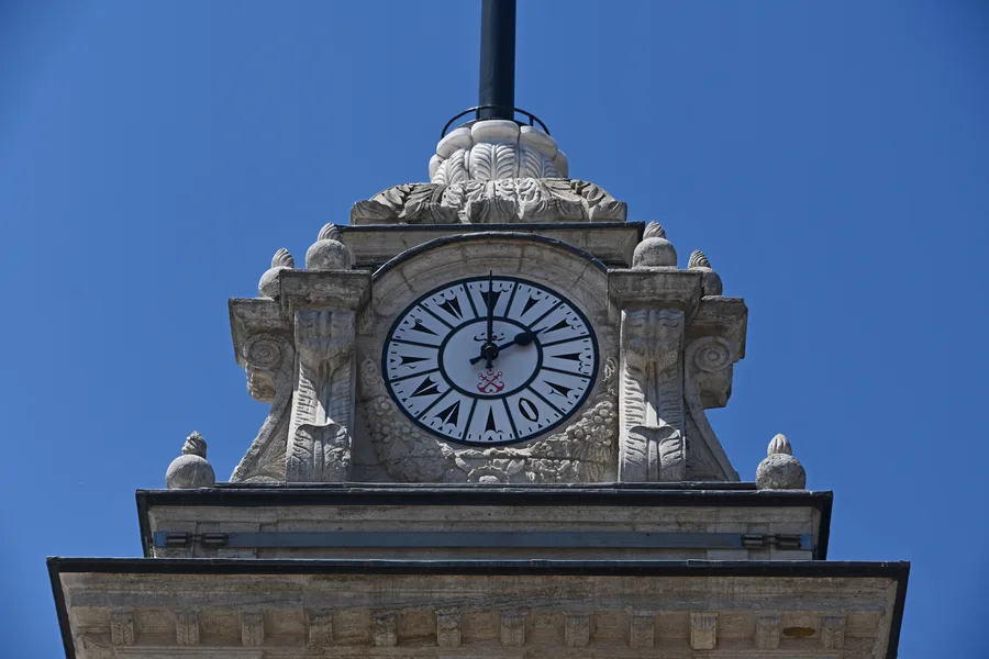 La parte superior de una torre del reloj de piedra contra un cielo azul intenso, con una esfera blanca con números romanos negros y adornos de piedra tallada a ambos lados