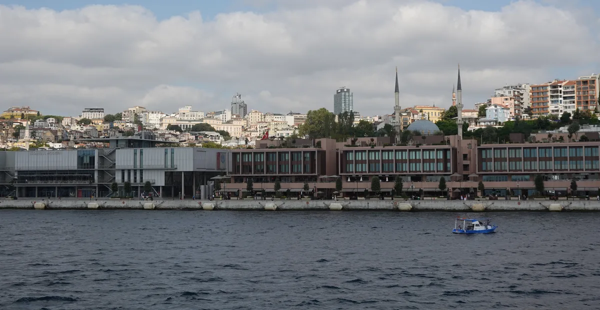 La larga fachada de un edificio moderno frente al mar, junto a un muelle de piedra en el Bósforo, con una pequeña embarcación azul en el agua y una ladera con casas y dos minaretes detrás