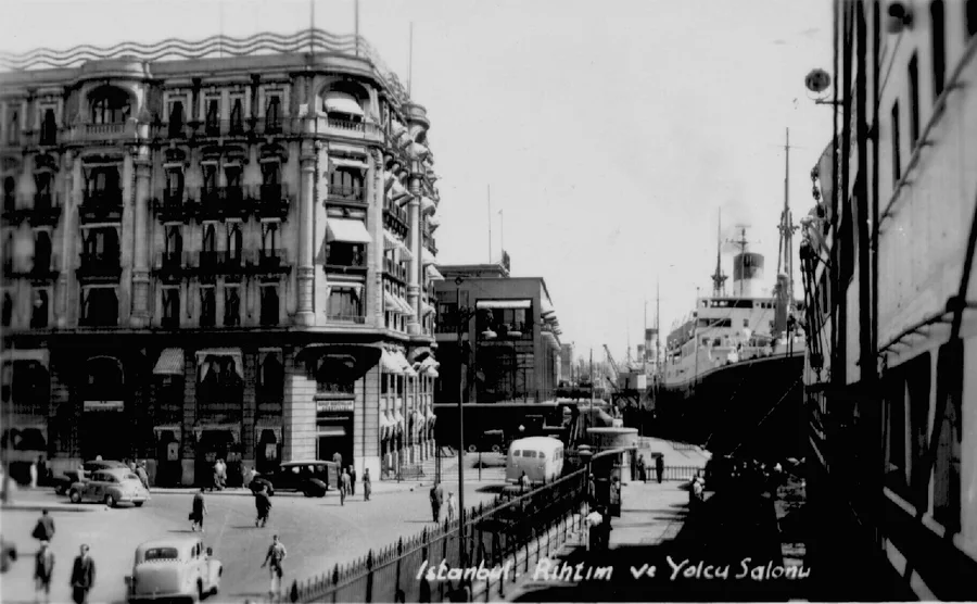 Una postal antigua en blanco y negro de una calle portuaria, con un gran edificio a la izquierda, coches y personas en la calzada y un buque de línea amarrado al muelle a la derecha