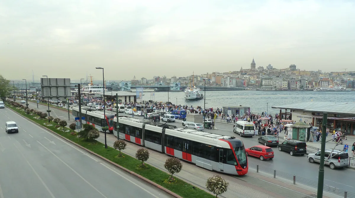 Un tranvía moderno rojo y blanco circula por una avenida junto al agua, con gente cerca de una parada y las colinas y torres de la orilla opuesta al fondo