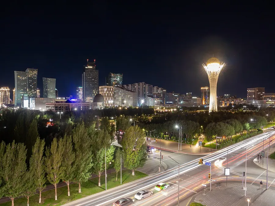 A tall white tower with a glowing top rises behind trees at night; a road with light trails runs below and tall buildings stand to the left