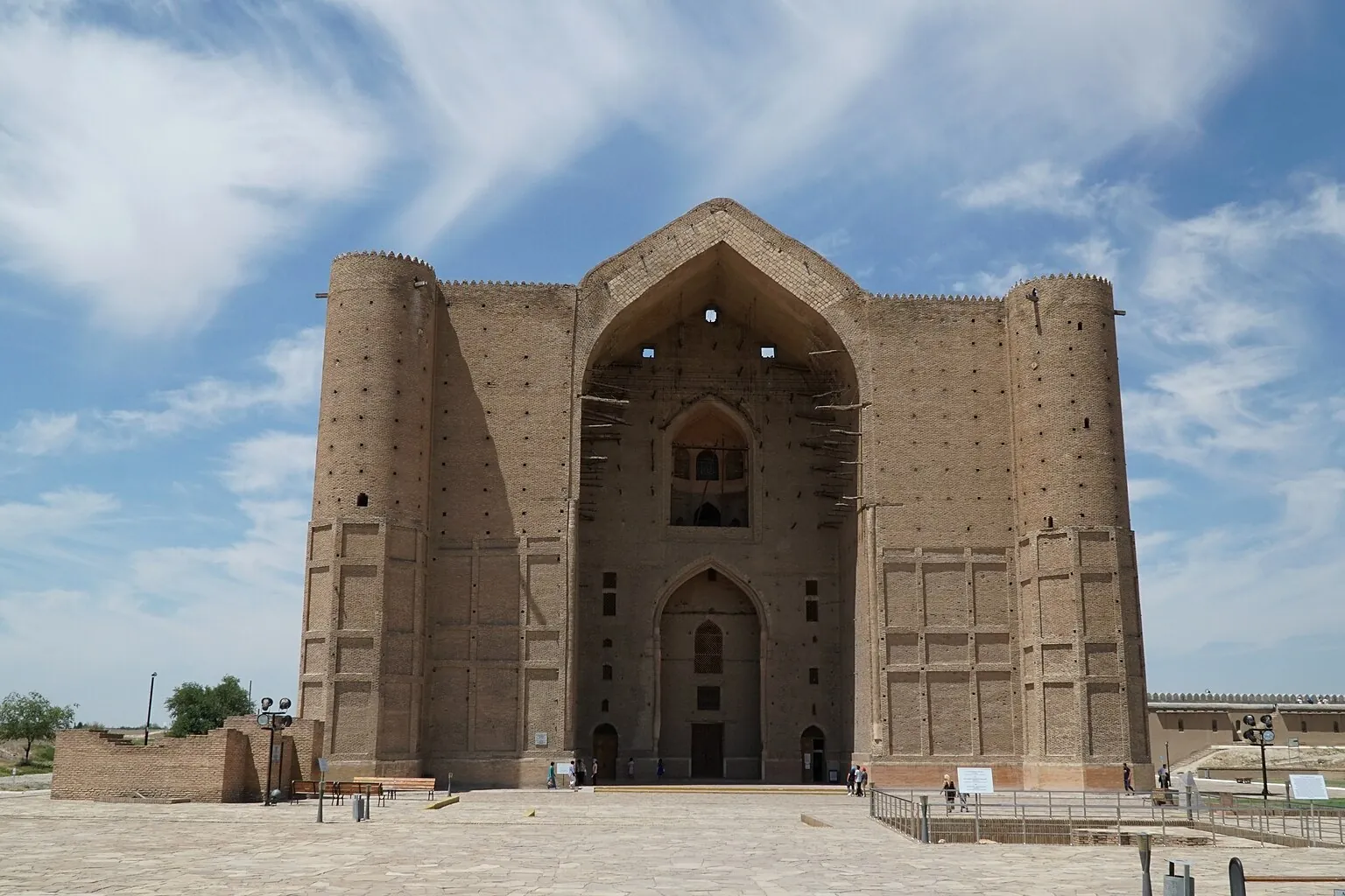 The front of a large brown brick mausoleum with a tall pointed-arch portal in the centre and a round tower on each side, seen across a paved square under a blue sky with thin clouds