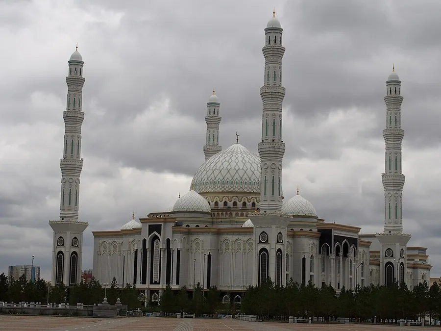 A white mosque with a pale dome, smaller domes and four tall minarets under a heavy grey sky