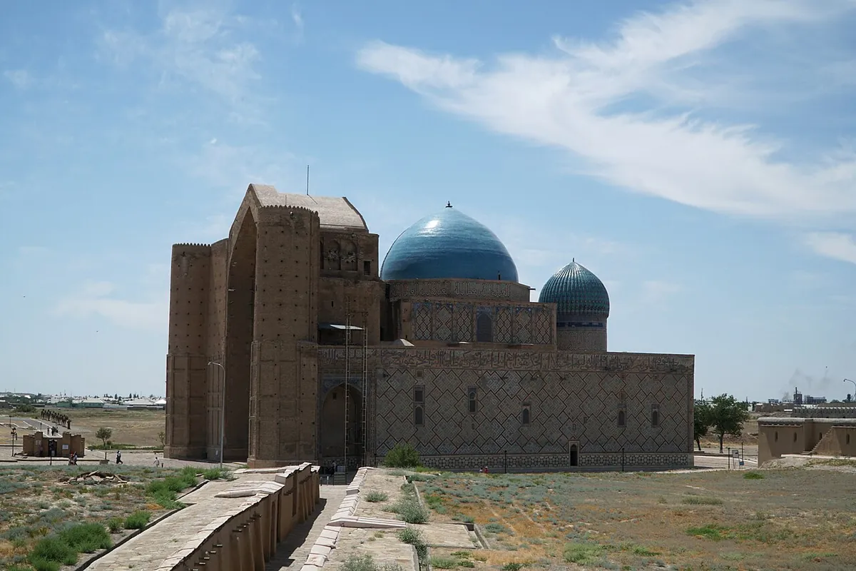 The side of the mausoleum with a blue ribbed dome and a smaller blue dome above a tile-patterned brick wall, and the tall portal at the left