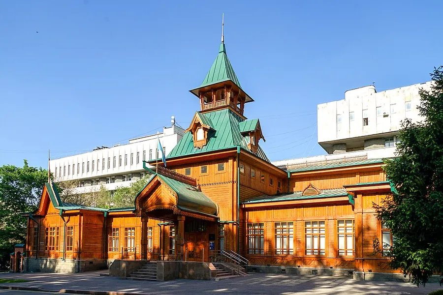 A carved wooden house with steep roofs, a small porch and a green steeple under a blue sky